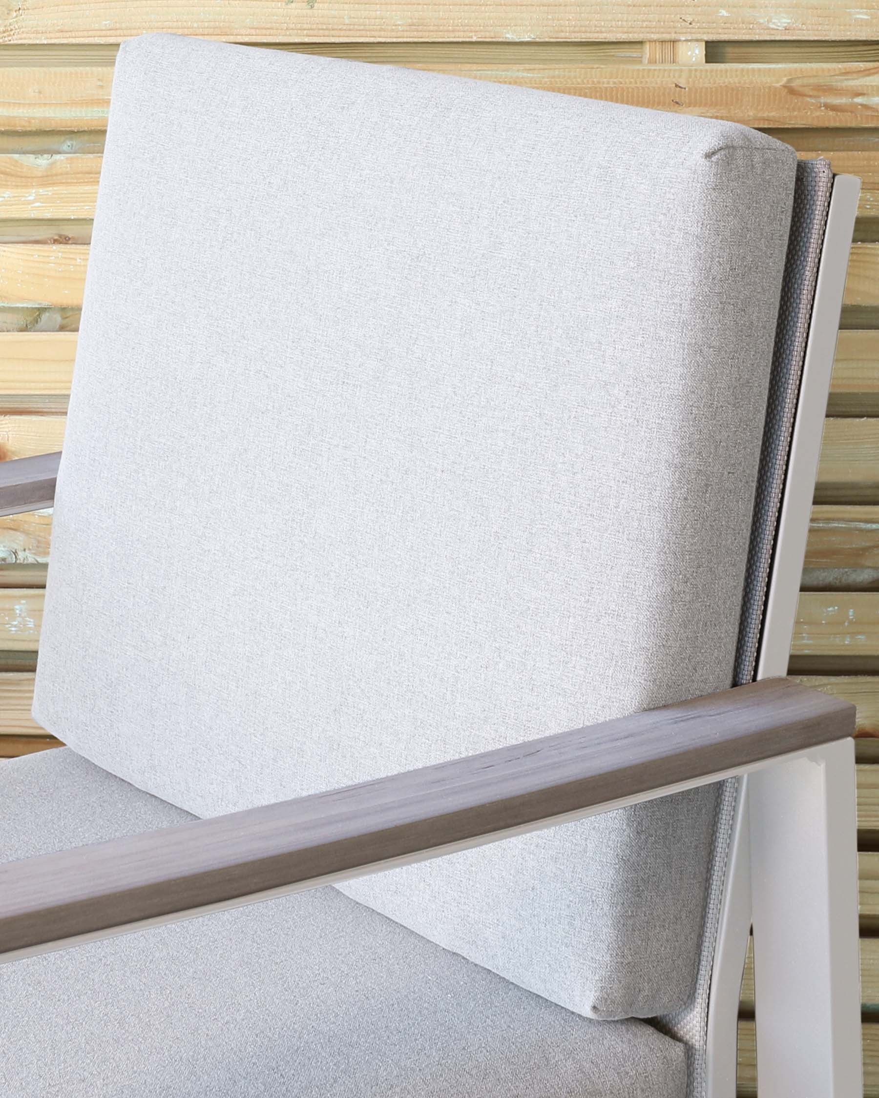 A close-up of a modern chair with a light grey upholstered back and seat, featuring sharp, clean lines and a minimalist design. The chair has a wooden armrest with a smooth finish that contrasts with the white painted frame, providing a contemporary look suitable for a variety of interior styles. The background shows a subtle, rustic wooden panelling that complements the chair's modern aesthetic.