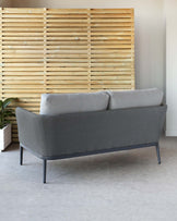 A modern, minimalist style grey fabric two-seater sofa with sleek black metal legs, positioned against a backdrop of a warm-toned wooden slat wall.