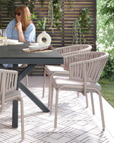 Modern outdoor furniture set featuring a large rectangular dining table with a dark grey top and a unique angular leg design in a darker tone. Surrounded by a collection of stylish chairs, each with a light beige fabric seat and a taupe, vertical rope backing that creates a contemporary airy look, all supported by slim, straight metallic legs.