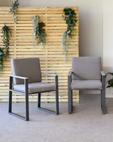 Two modern armchairs with grey upholstery and sleek, matte black metal frames, set against a minimalist background with a wood-slat wall adorned with cascading greenery.