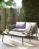 Modern outdoor furniture featuring a sleek grey two-seater sofa with clean lines and light grey fabric cushions, paired with a rectangular, minimalist white coffee table with slender metal legs. The set creates a serene and stylish ambiance on a patio surrounded by greenery.