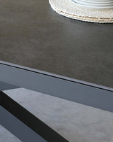 Modern minimalist table with a dark grey matte finish tabletop and a sleek black angular metal base, shown with a woven placemat and ceramic dish on top, against a grey flooring background.