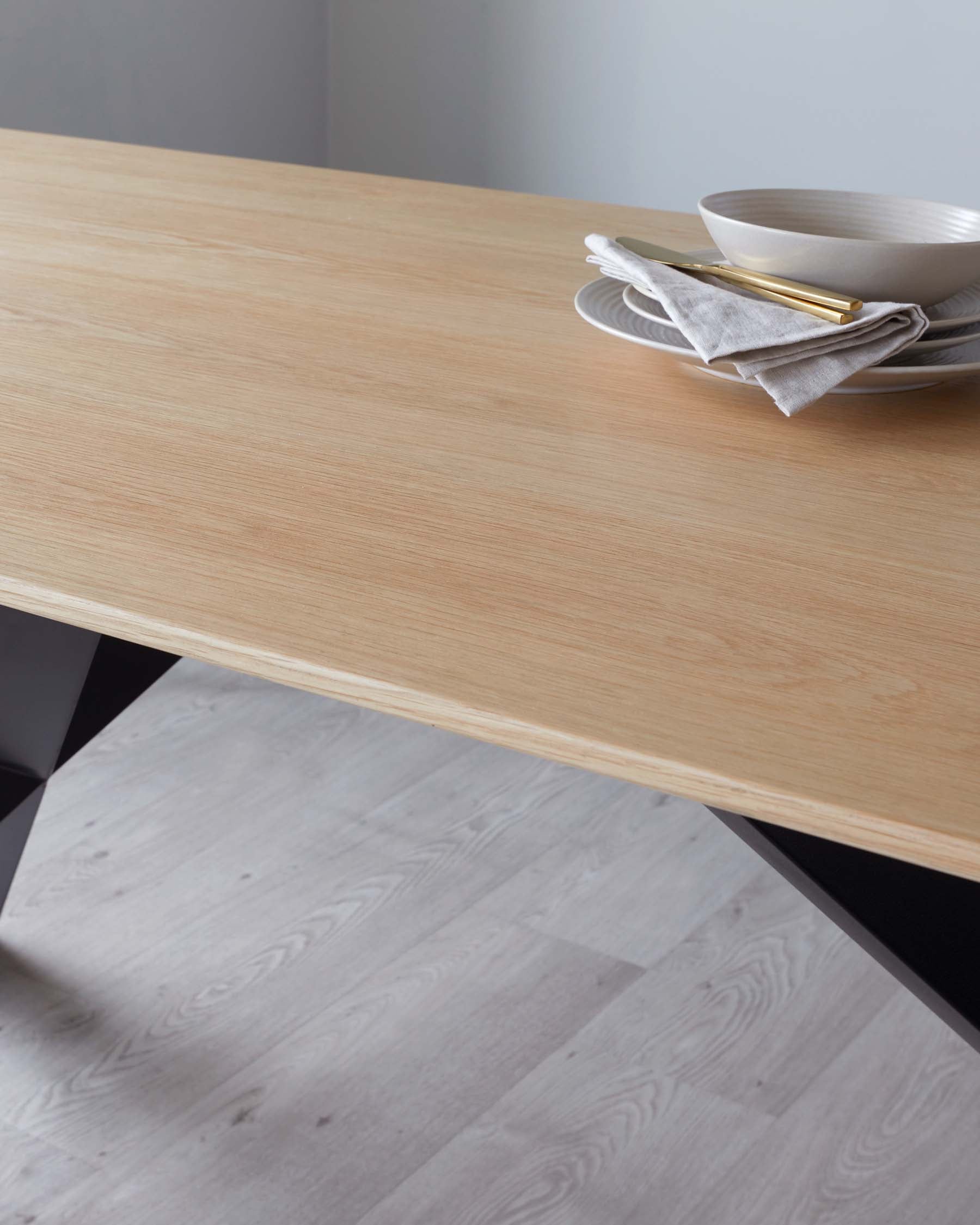 Modern minimalist wooden dining table with a smooth, light wood finish, featuring sleek, angular black legs. The table is partially set with a stack of ceramic dishes and a set of gold cutlery wrapped in a grey linen napkin, indicating a refined yet simplistic aesthetic.