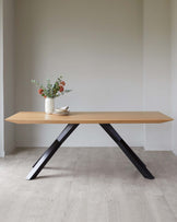 Modern minimalist wooden table with a natural finish and angled black metal legs, accompanied by a small white ceramic vase with flowers and a stack of dishes on top, set against a neutral wall and light wooden flooring.