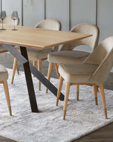 Modern dining room furniture set featuring a rectangular wooden table with contrasting black metal angled legs, complemented by four plush velvet upholstered chairs in a light taupe colour with wooden legs accented with gold metal tips. The set is arranged on a textured white area rug.