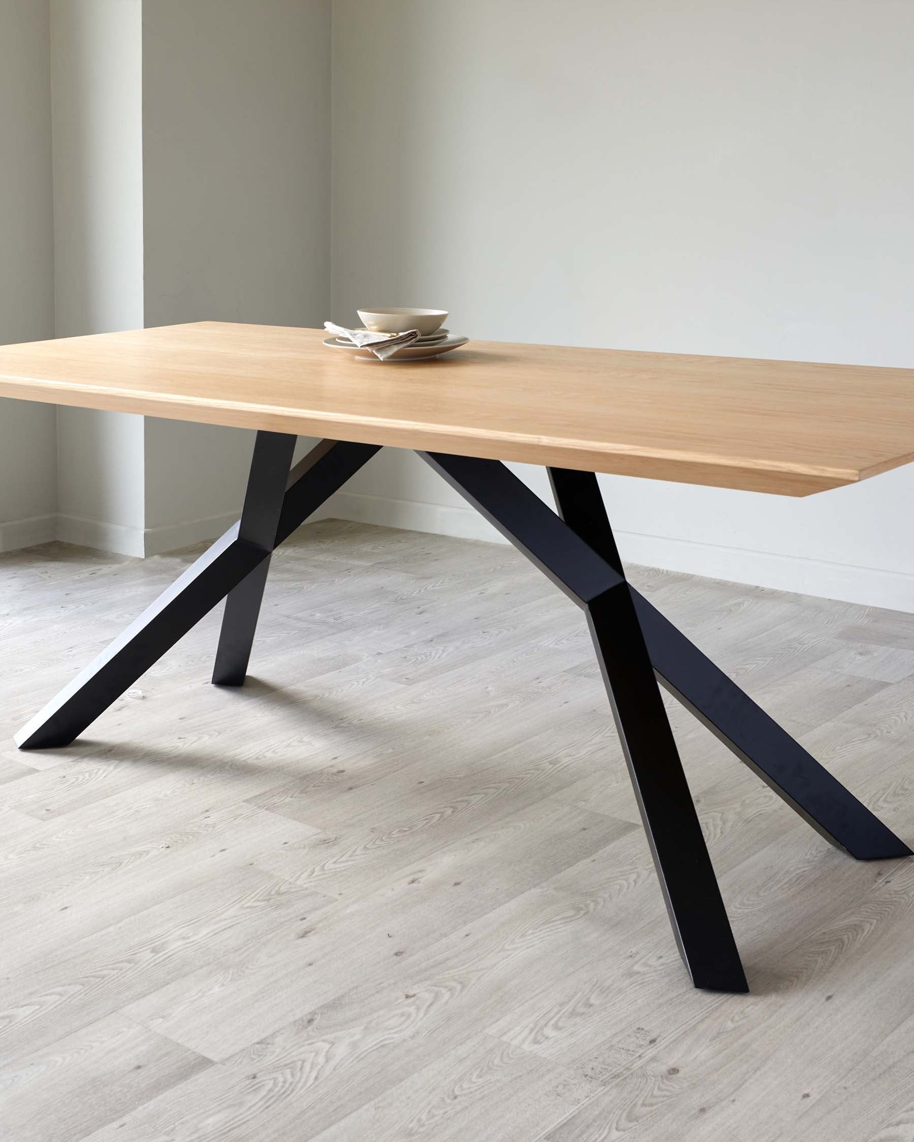 A modern minimalist dining table with a smooth, light wood tabletop and angular, matte black metal legs in an X-shape. The table is set against a neutral backdrop and there are stacked white bowls on the tabletop, adding a touch of elegance to the simple design.