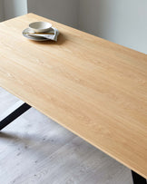 Modern minimalist wooden dining table with a natural oak finish and black metal legs, accompanied by a small stack of white ceramic plates and a folded napkin on top.