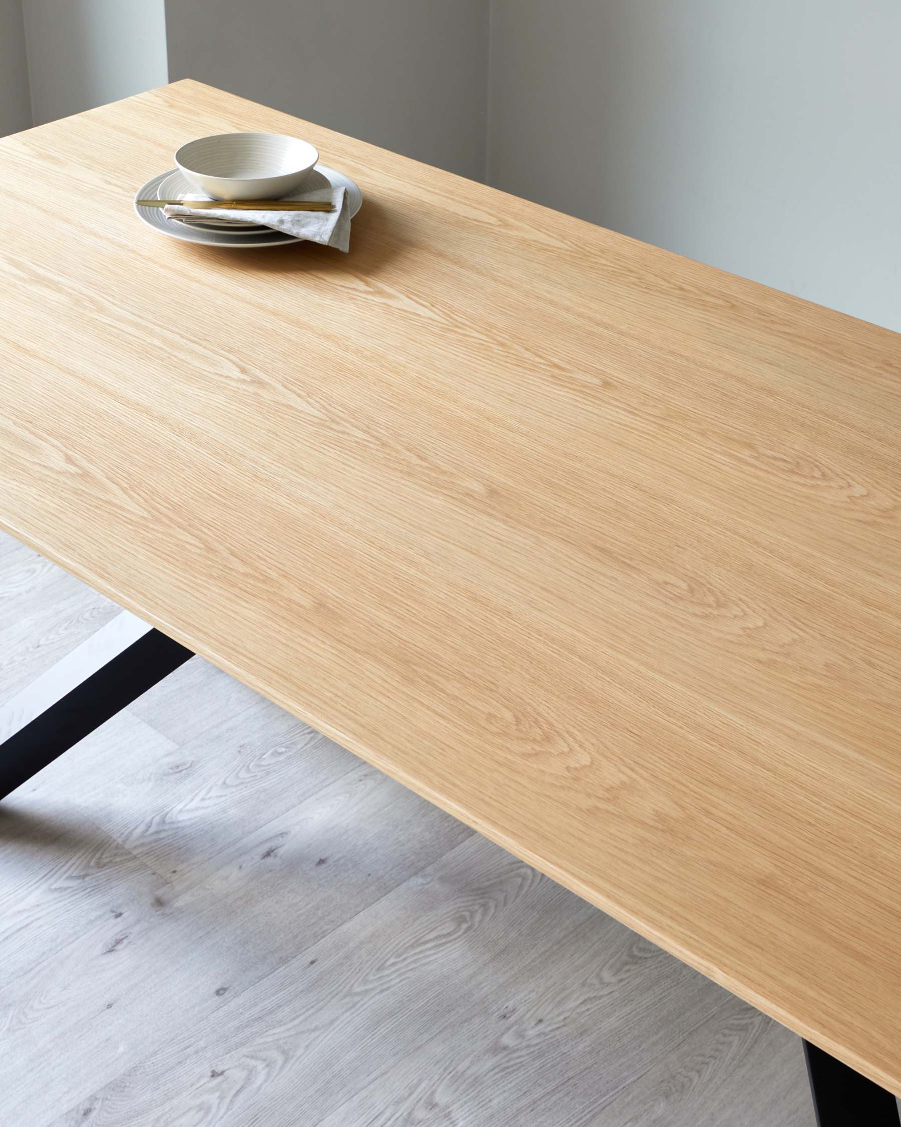 Modern minimalist wooden dining table with a natural oak finish and black metal legs, accompanied by a small stack of white ceramic plates and a folded napkin on top.