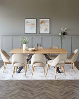 A modern dining set featuring a rectangular wooden table with sleek black metal legs, surrounded by eight upholstered chairs in a light taupe fabric with a smooth, rounded back design and angled wooden legs. The set is staged on a textured grey and white area rug, and the scene includes simple decorative elements such as framed wall art, green foliage in vases, and minimalist tableware.