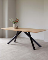 A modern rectangular dining table with a light natural wood tabletop and a distinctive black geometric metal base, set in a neutral-toned room with a vase of flowers and a small stack of plates as decor.