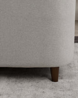 Close-up view of a contemporary sofa with a textured light grey fabric upholstery and a single cylindrical dark wooden leg visible. The image showcases the clean lines and modern aesthetic of the design.