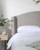 Elegant modern bedroom with a curved light grey fabric headboard, white bedding set, and a minimalist light wooden bedside table featuring a simple white ceramic cup. A decorative green potted plant and dried wheat stalks add natural elements to the serene space.