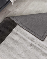 Contemporary area rugs in contrasting shades, featuring a dual-layered design with one rug partially overlapping the other. The bottom rug is in a light grey tone with a subtle texture, while the top rug boasts a darker, charcoal grey hue with a fine weave detail and refined border stitching. Both rugs lay on a light wooden floor, complementing a modern aesthetic.