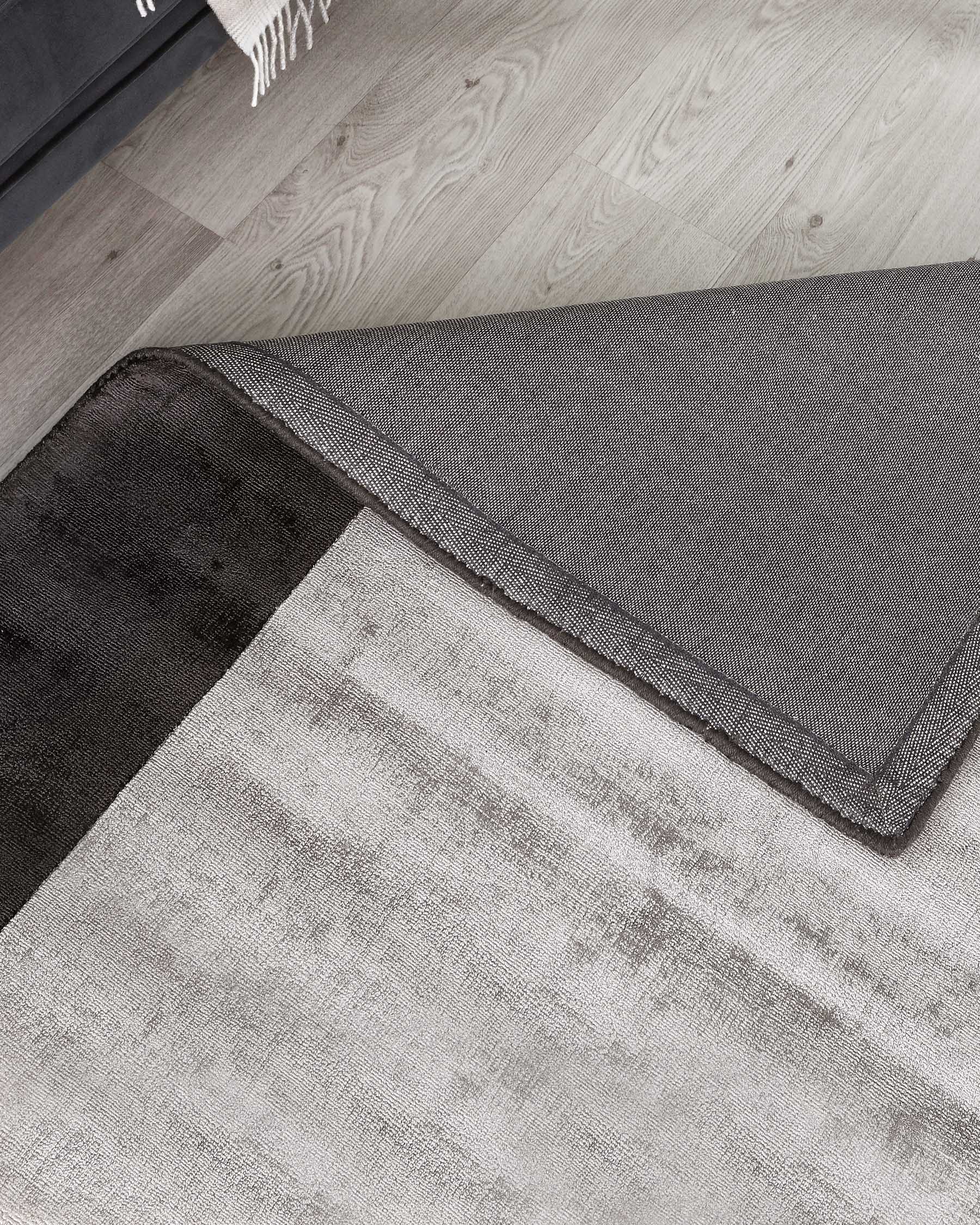 Contemporary area rugs in contrasting shades, featuring a dual-layered design with one rug partially overlapping the other. The bottom rug is in a light grey tone with a subtle texture, while the top rug boasts a darker, charcoal grey hue with a fine weave detail and refined border stitching. Both rugs lay on a light wooden floor, complementing a modern aesthetic.