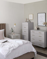 A contemporary bedroom suite featuring a taupe upholstered bed flanked by a small grey nightstand on the left, and a taller, matching grey drawer chest on the right. Across from the bed stands a larger grey dresser with six drawers, all pieces accented with sleek golden handles.