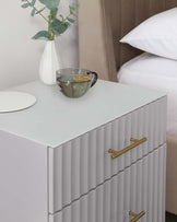 A contemporary white bedside table with a ribbed texture on the drawers and elegant gold handles, positioned next to a bed with white linen. The tabletop holds a vase with greenery, a clear glass cup with a hot beverage, and a white round object, possibly a coaster or decorative item.