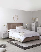 Upholstered platform bed with a channel-tufted headboard in a neutral taupe, flanked by two white modern nightstands with gold accents, and a matching white dresser with gold handles set against a light grey wall. A decorative round gold mirror hangs above the bed.