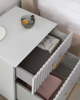 Modern white bedside table with an open drawer revealing organized compartments, some filled with neatly folded fabrics. The drawer features unique wave-textured fronts and elegant golden handles. A simple, sleek design with a glossy finish, showcased next to a soft-toned bed. A clear glass bowl with a slice of orange and a white round plate are placed on top of the table, adding a pop of colour and a decorative touch.