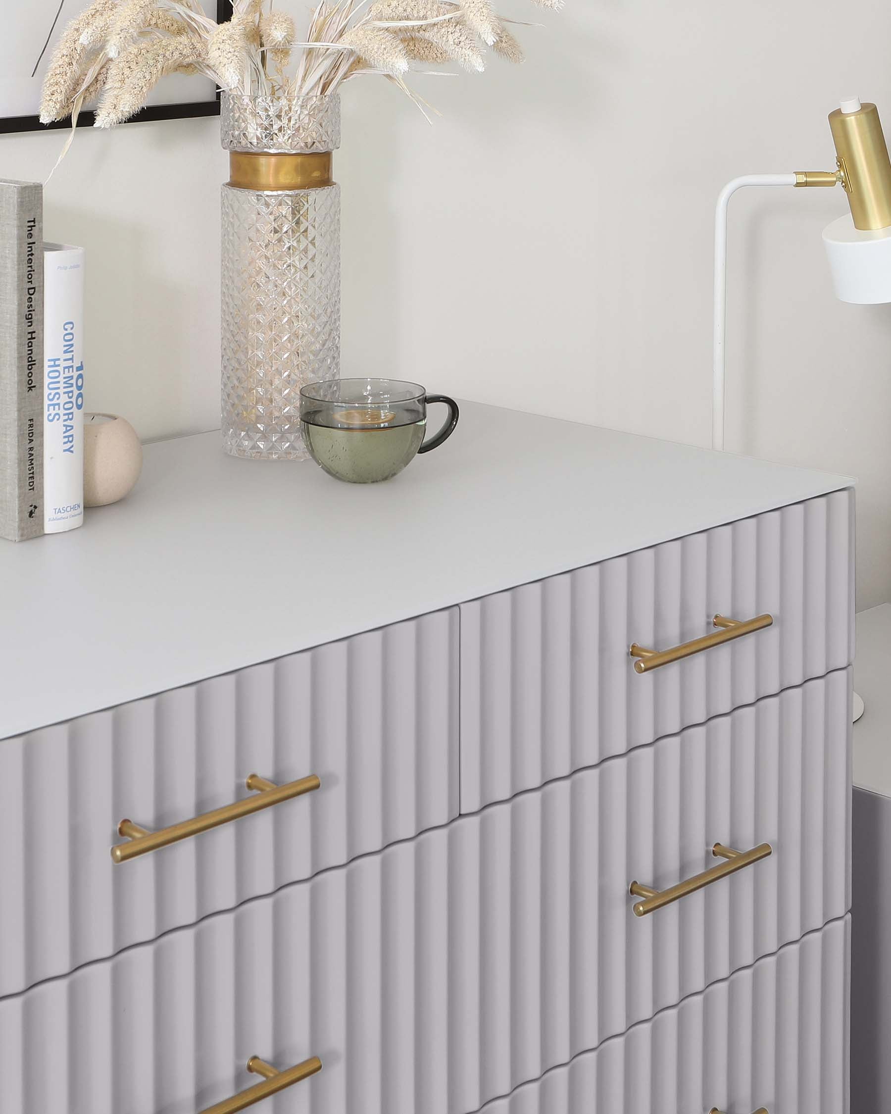 Modern light grey dresser with textured front and brass handles. The top surface is adorned with a decorative vase, a book, and a clear glass cup. A white wall-mounted lamp with a gold accent is positioned above the dresser.