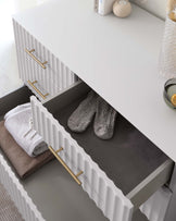 White modern dresser with ribbed drawer fronts and brass handles, partially open to reveal folded towels and slippers inside.