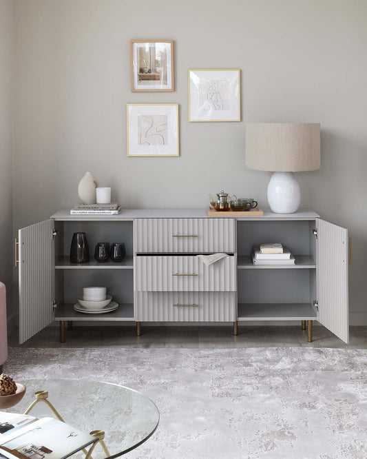 A contemporary sideboard in a soft grey finish features fluted front panels and metallic horizontal handles. The unit has two doors and three central drawers, with angled wooden legs. Above it, a round white lamp with a textured base and a beige lampshade complements the sideboard. On the left, a round glass coffee table with a gold rim and legs is visible. The floor is covered with a distressed grey area rug, and the walls are adorned with framed artwork.