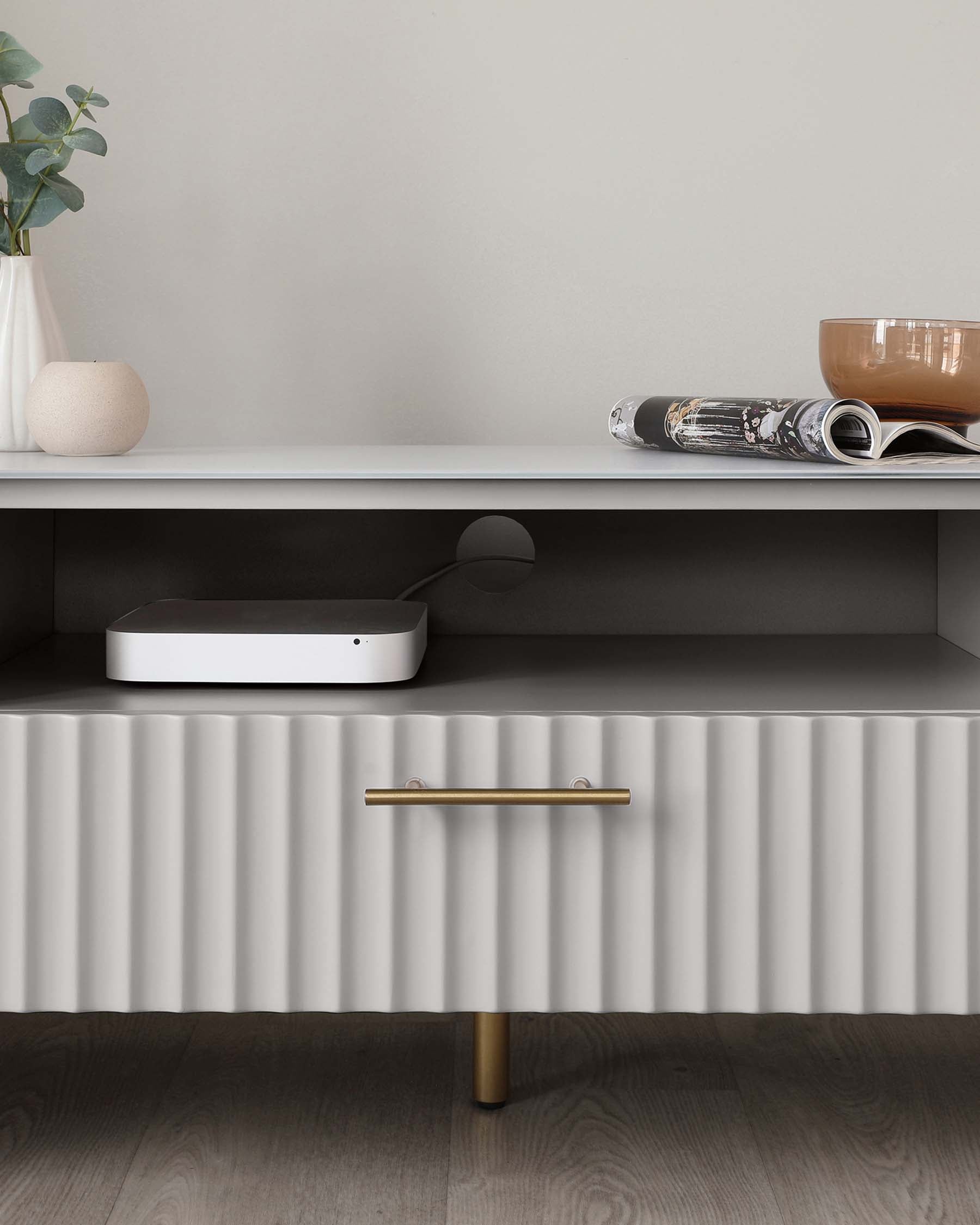 A modern minimalist TV stand or media console in grey with a smooth top shelf and grooved lower cabinet façade. It features a single cylindrical wooden leg supporting one side, while the other is flush with the wall. The cabinet door sports a sleek, metallic horizontal pull handle. On the top shelf, there is a ceramic vase with greenery, a large round candle, and a rolled-up magazine, creating a stylish and contemporary look.