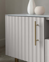 Modern fluted-front sideboard in white with brass-tone metal handles and slender, tapered legs, accompanied by a ceramic vase and stone sphere as tabletop decor.