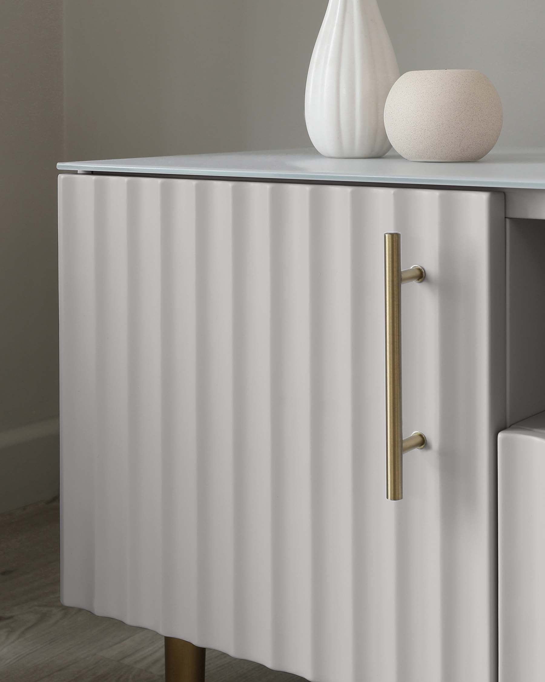 Modern fluted-front sideboard in white with brass-tone metal handles and slender, tapered legs, accompanied by a ceramic vase and stone sphere as tabletop decor.