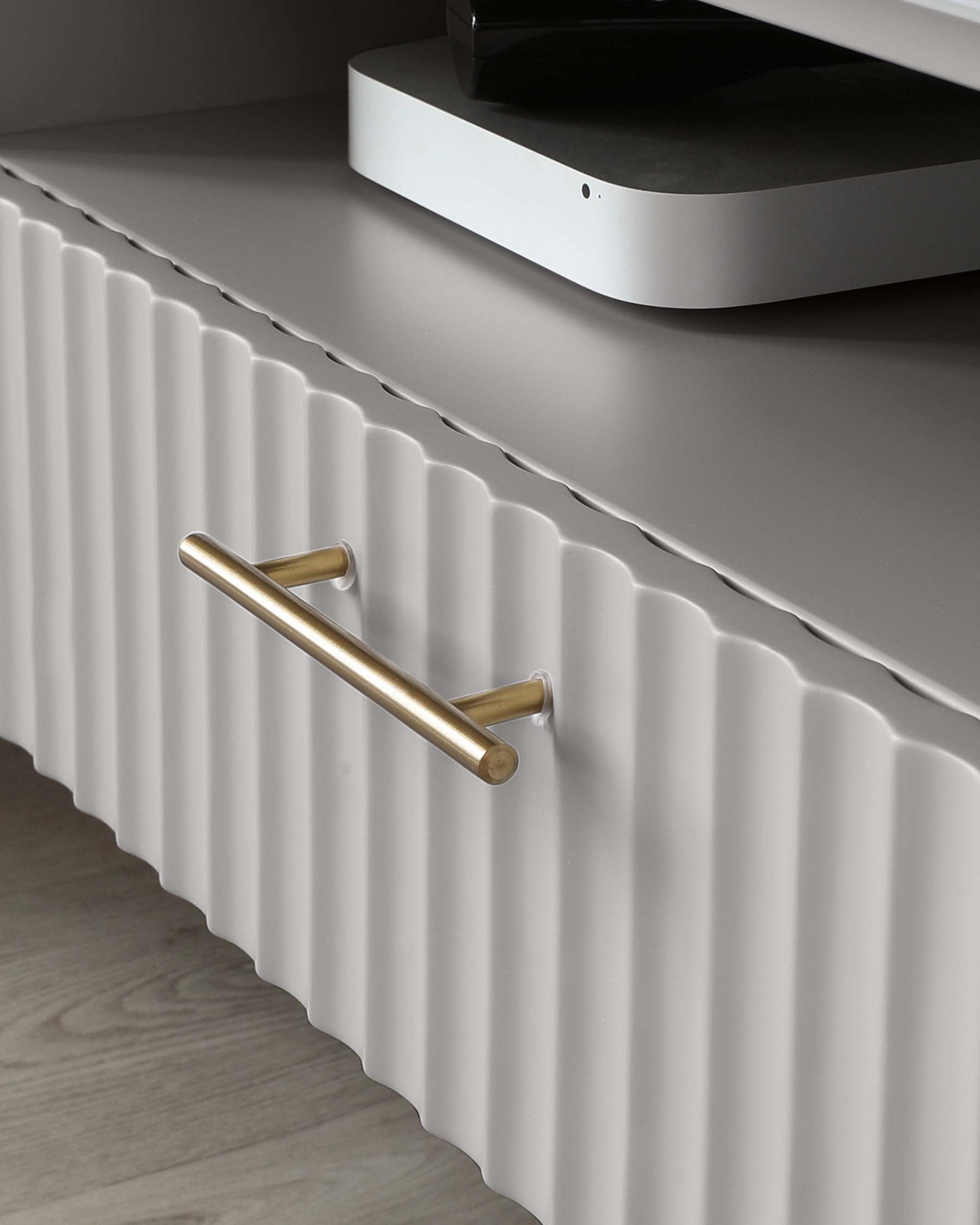 Contemporary white cabinet with textured front design and brushed gold horizontal handle; a modern electronic device is placed on top of it.