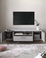 Modern white TV stand with ribbed detailing and gold-finished handles, featuring multiple open shelves and closed cabinets on an oak-toned base, styled with a minimalistic vase and books. A muted patterned rug lies beneath, complementing the contemporary aesthetic.