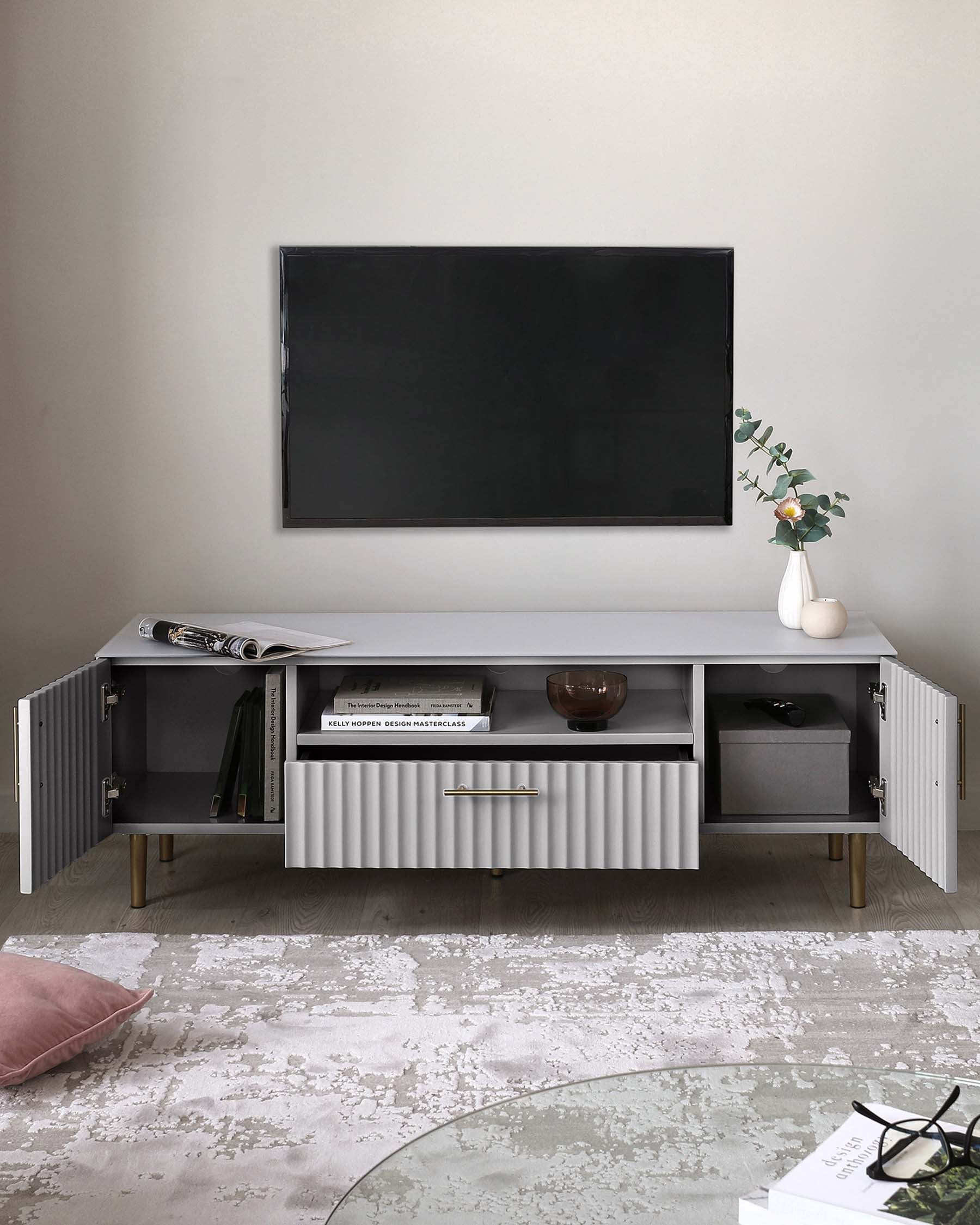 Modern white TV stand with ribbed detailing and gold-finished handles, featuring multiple open shelves and closed cabinets on an oak-toned base, styled with a minimalistic vase and books. A muted patterned rug lies beneath, complementing the contemporary aesthetic.