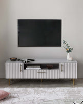 A contemporary white sideboard with a textured front, gold handles, and tapered wooden legs. The sideboard has two cabinet doors and a central shelf housing decorative items, positioned below a wall-mounted television, flanked by ornamental plant and books. A patterned light grey and white area rug is partially visible beneath it.