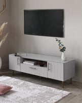 Modern minimalist TV stand in light grey with ribbed front panels and brass-finished handles and legs, featuring two cabinets and a central open shelf with a decorative vase and books.