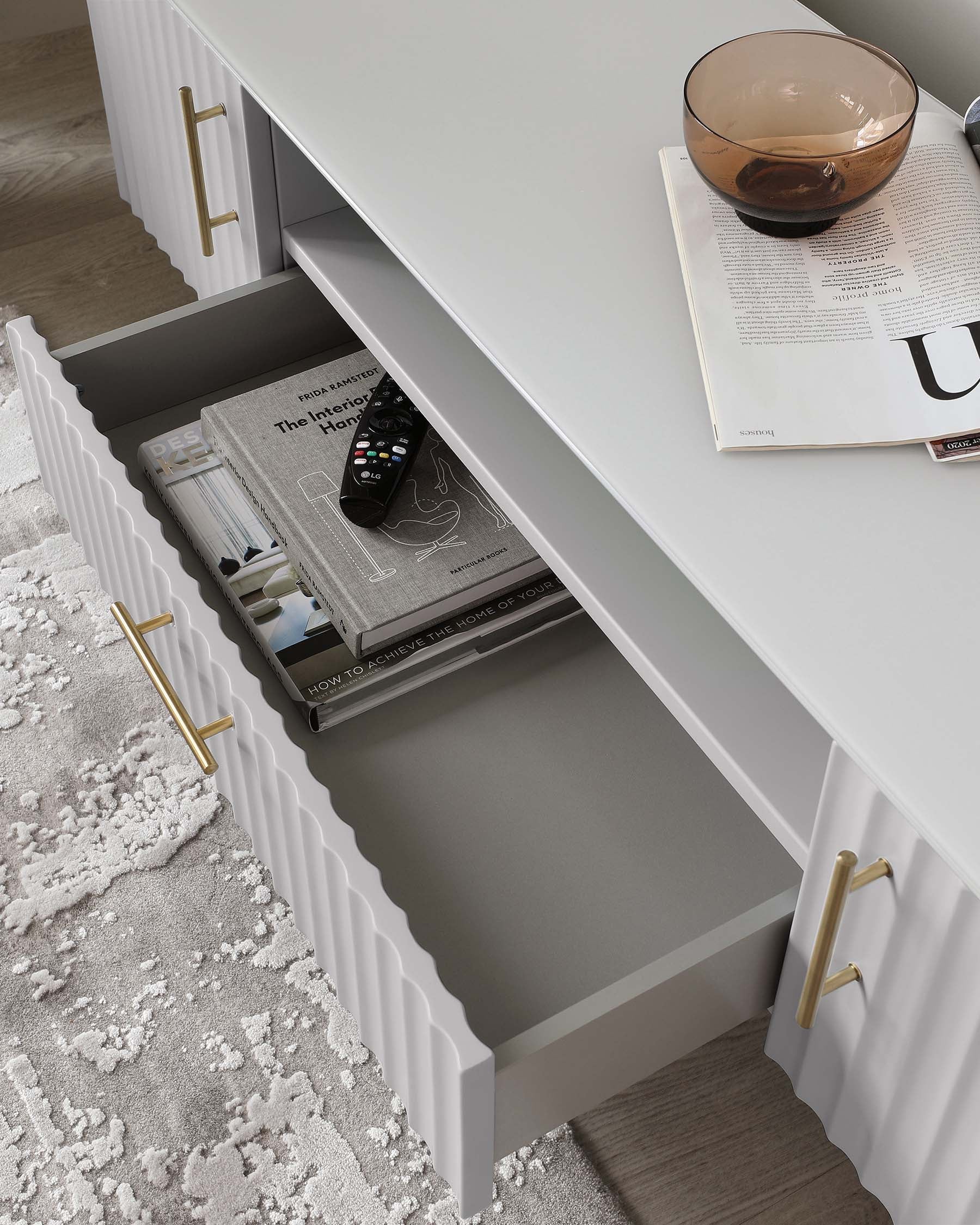 A modern white console table with fluted drawer detail and brass handles, partially open to reveal magazines and a remote control, with a decorative bowl on top beside an open magazine. The console rests on a plush grey and white patterned area rug.