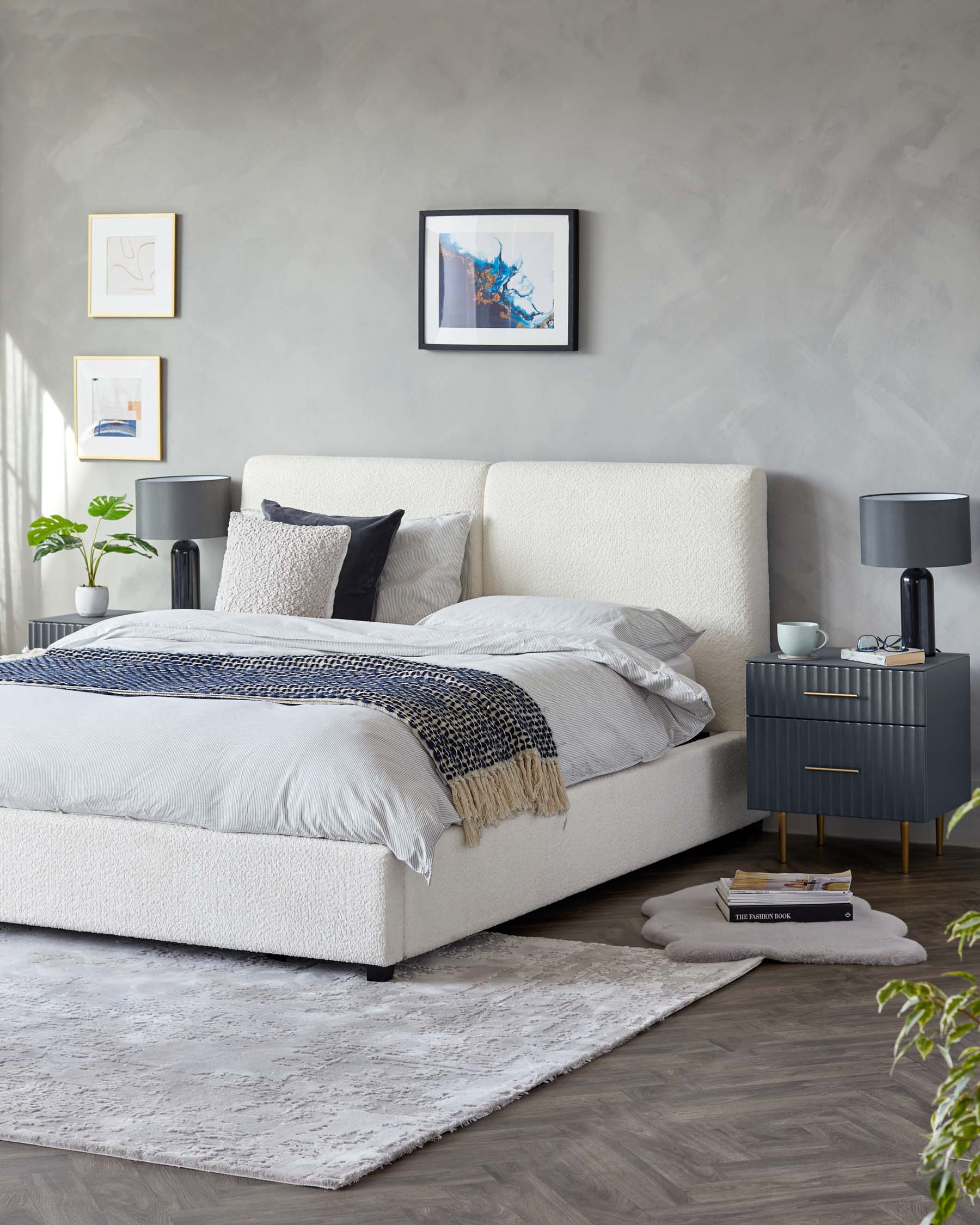 Contemporary bedroom setup with a plush upholstered bed frame in light cream fabric, complemented by grey textured bedding and assorted throw pillows. Beside the bed stands a modern charcoal grey nightstand with brass accents, featuring drawers for storage. The room is anchored by a soft textured grey area rug, which adds a layer of warmth and comfort to the sleek hardwood flooring.