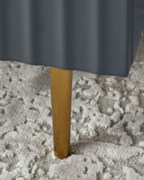 Close-up view of a furniture leg with a natural wood finish, set against a soft, plush grey carpet. The furniture piece has a dark grey fabric covering draped above the leg, suggesting a modern and elegant design approach.