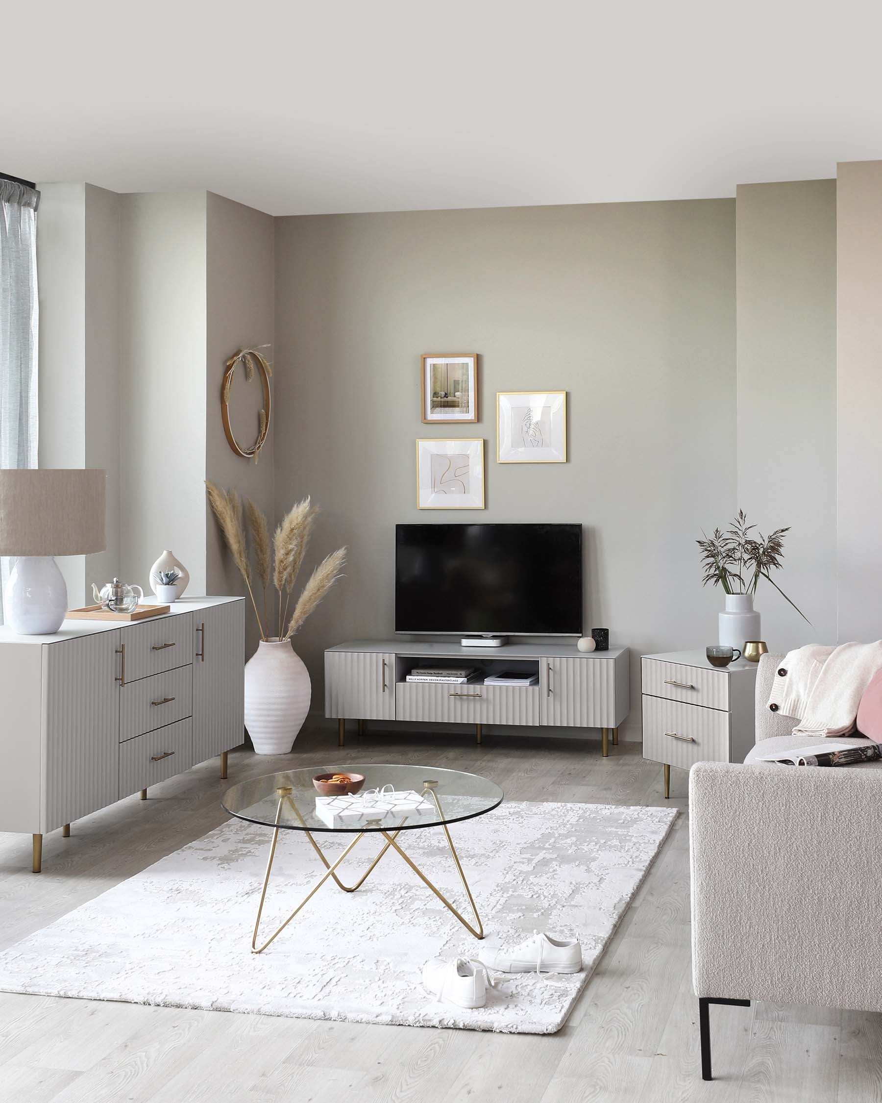 A modern living room featuring a glass coffee table with a gold frame, two off-white side cabinets with brass handles, a matching TV stand with ribbed doors, and a light grey upholstered armchair with black legs. A soft texture area rug lies beneath the coffee table.