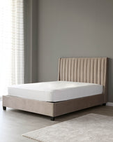 Modern upholstered bed frame with tufted headboard in a neutral taupe colour, and a white mattress set against a muted grey wall next to a translucent-curtained window, with a light grey area rug partially visible on a light wood floor.