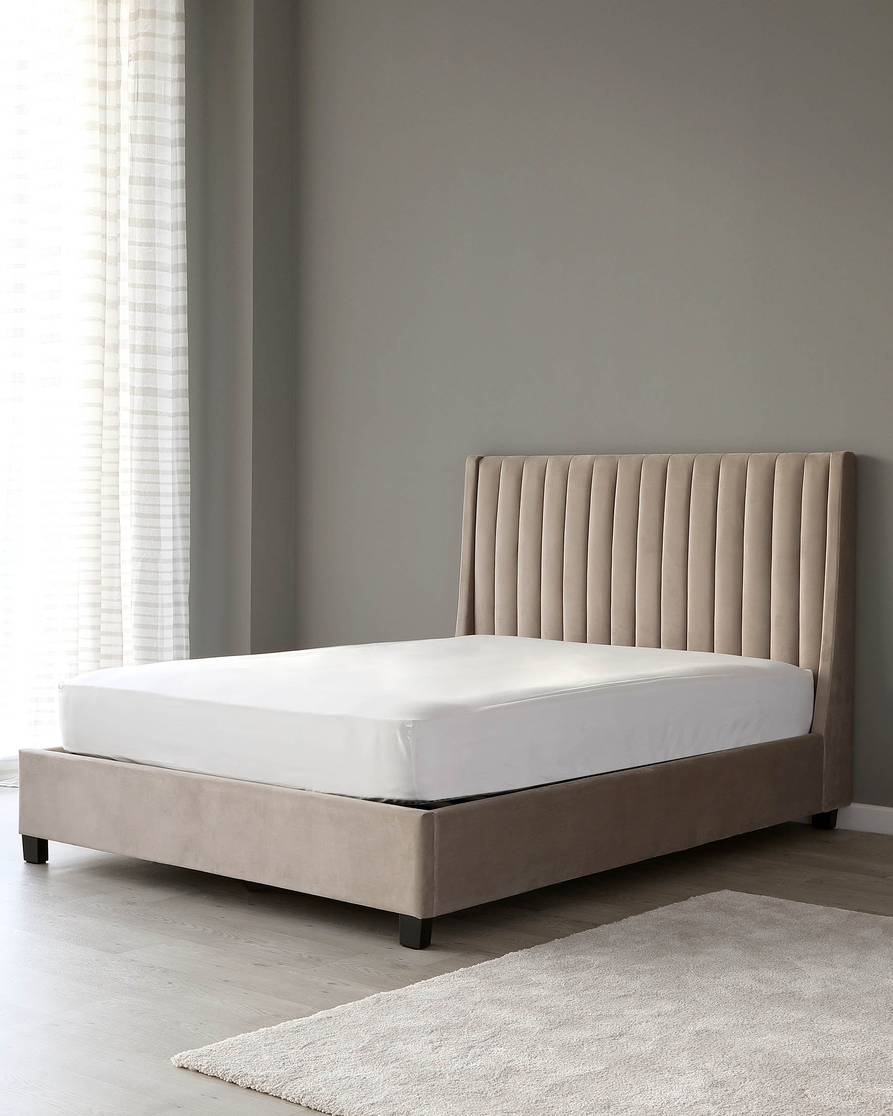 Modern minimalist style queen-sized bed with an upholstered headboard featuring vertical channel tufting, set on a low-profile platform frame with dark wooden feet, complete with an unadorned white mattress. The bed is positioned in a room with light grey walls and sheer curtains, accompanied by a soft grey area rug on the floor.