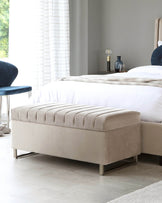 Elegant modern bedroom furniture set including a beige upholstered bed with a large headboard and white bedding, complemented by a matching beige upholstered bench at the foot of the bed. The bench features tufted detailing and stands on slender metallic legs. A small side table with sleek lines, housing a contemporary lamp and a blue vase, sits beside the bed on a light wood floor, with sheer curtains allowing soft, natural light into the room.