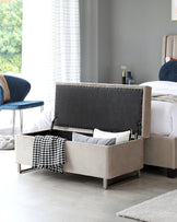 Beige upholstered storage bench with a tufted grey interior, situated at the foot of a bed, with metallic legs and a houndstooth throw blanket draped over one corner.