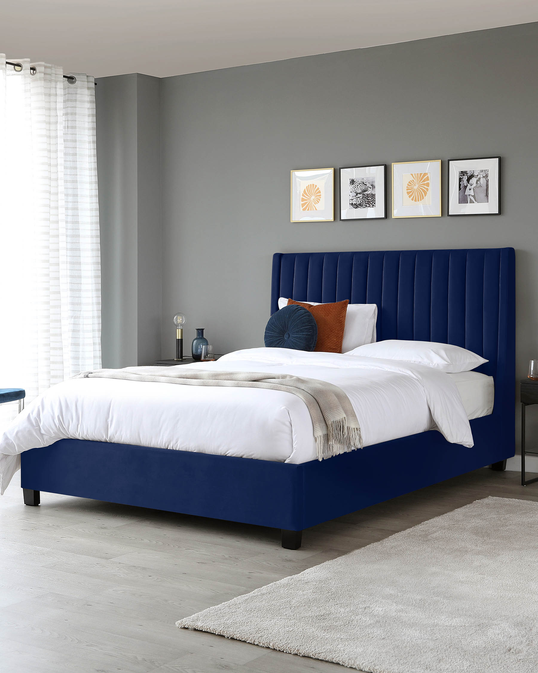 Navy upholstered bed with vertical stitching, white bedding, decorative pillows, a nightstand, and a soft area rug.