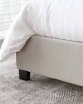 Modern minimalist bed frame with a light grey upholstery and squared black legs, partially covered by a crisp white duvet. The bed stands on a textured beige carpet.