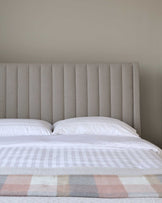 Upholstered headboard in a neutral fabric with vertical channel tufting detail, paired with a bed dressed in crisp white linens and a striped bedspread with soft pastel squares at the foot.