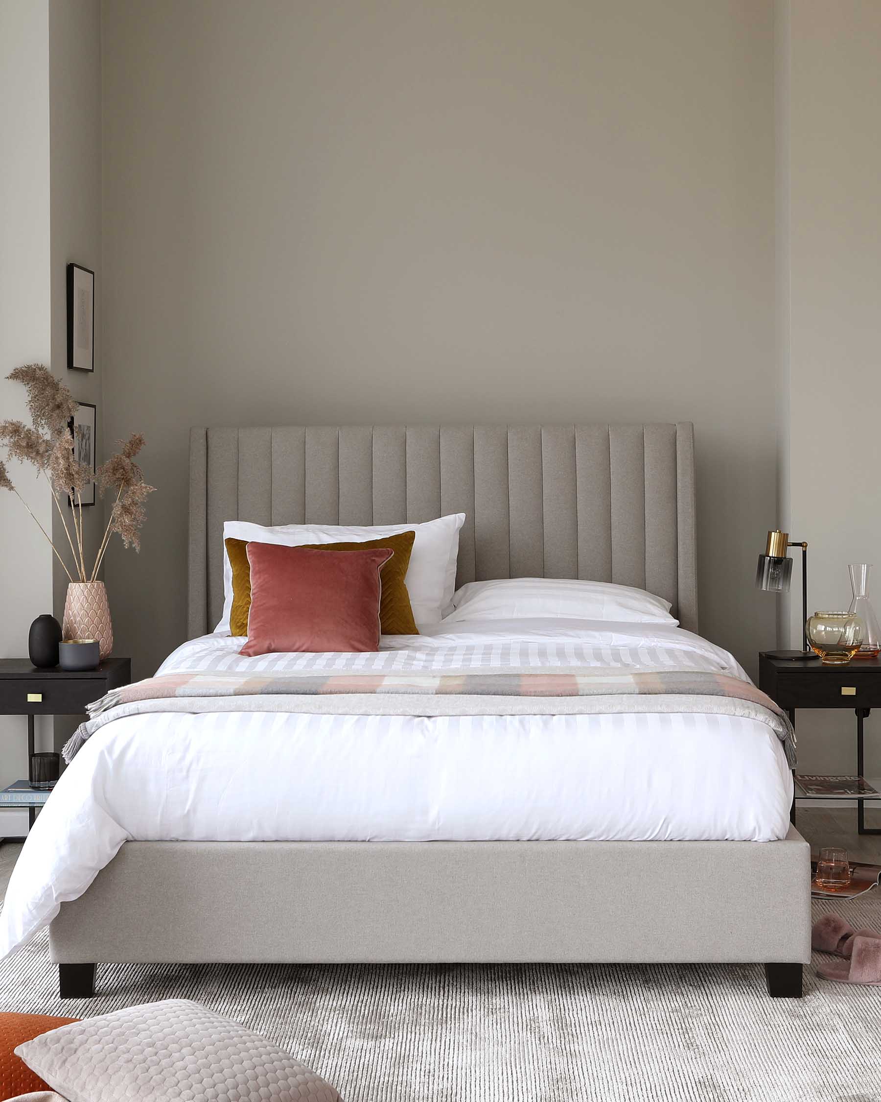 Modern bedroom featuring a grey upholstered platform bed with a tall headboard and solid wood dark-stained legs, complemented by white and plaid bedding with accent pillows in mustard and deep red. Beside the bed is a black nightstand with a lower shelf, displaying books, a table lamp, and decorative items.