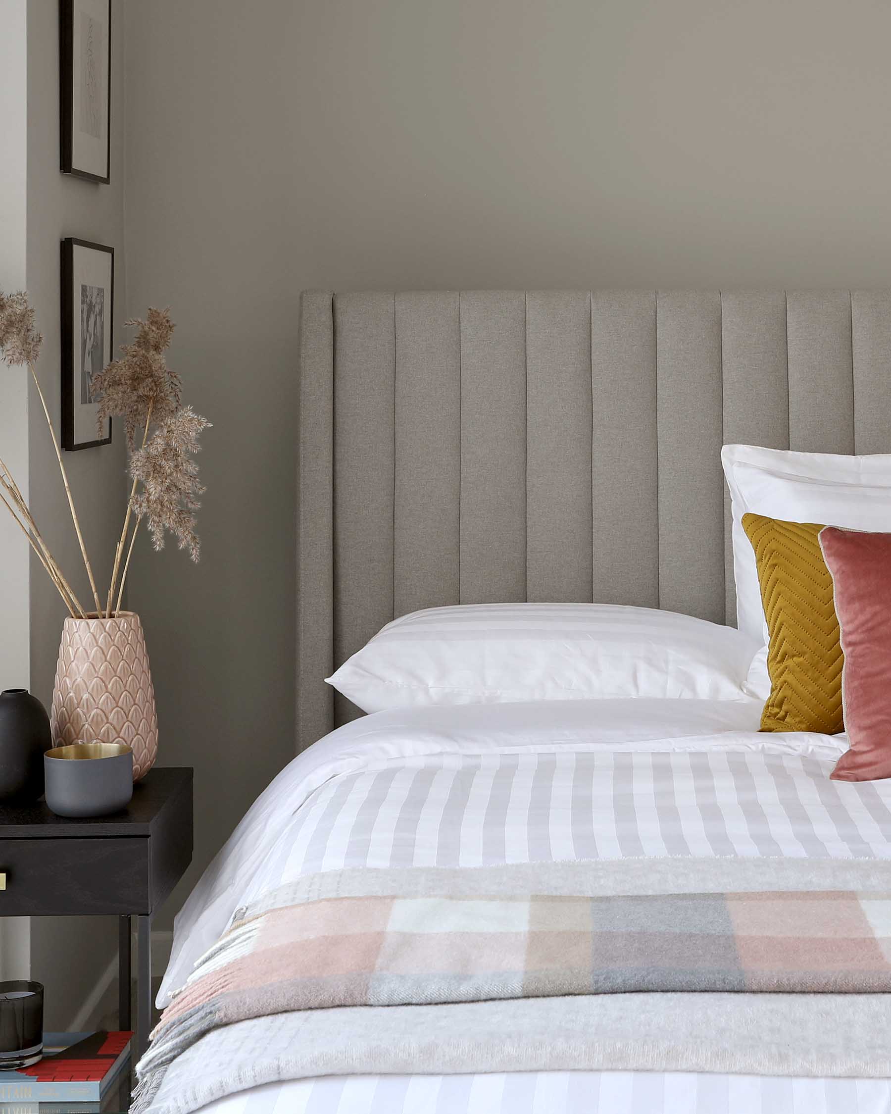 Contemporary bedroom featuring a large, upholstered grey headboard with vertical panelling, flanked by a sleek black nightstand with a textured pink vase and decorative bowl. The bed is dressed with striped and solid pillows and a striped comforter in pastel hues atop crisp white linens.