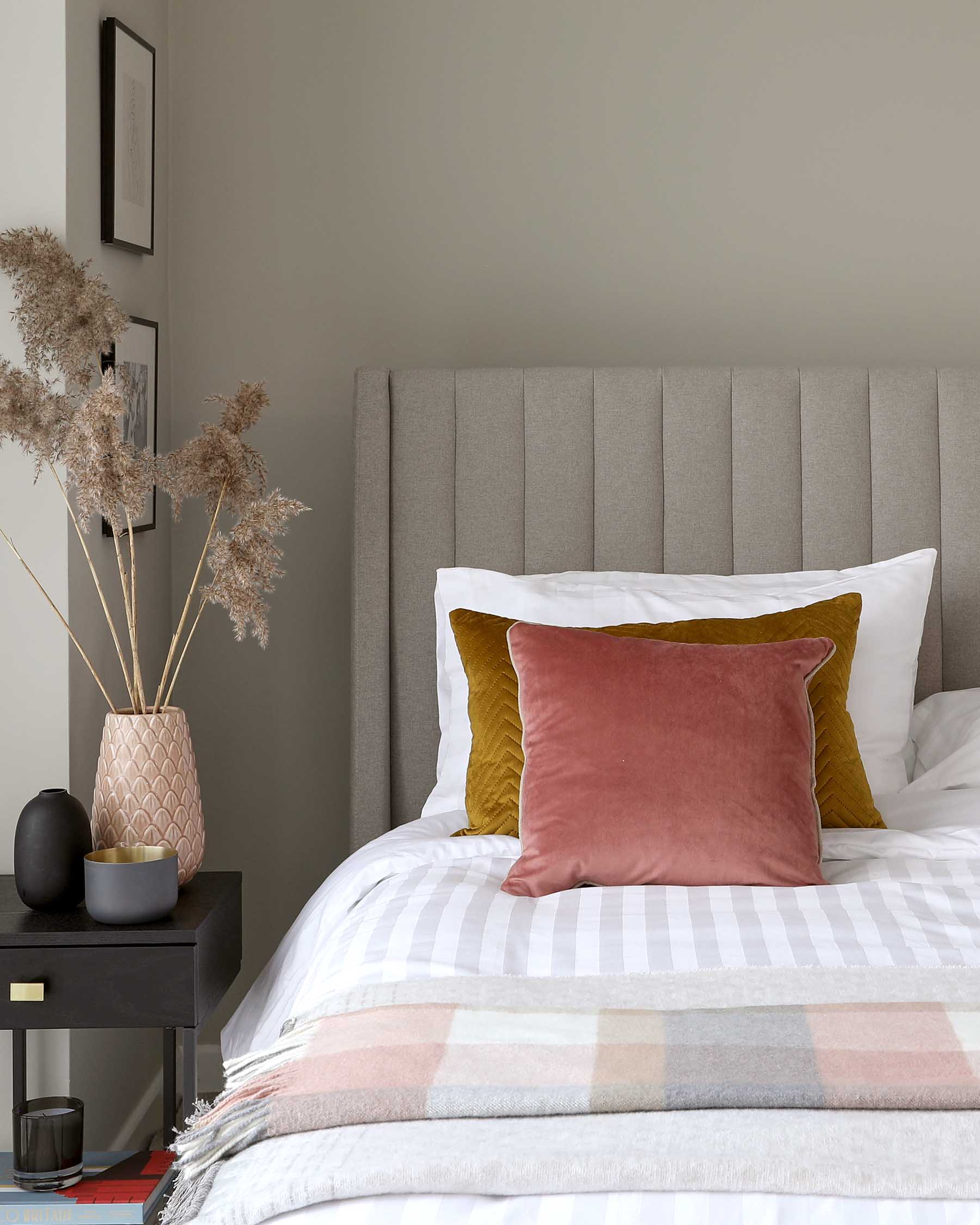 Elegant bedroom scene featuring a minimalist black bedside table with a brass knob, and a plush grey upholstered bedhead with vertical stitch line patterning.