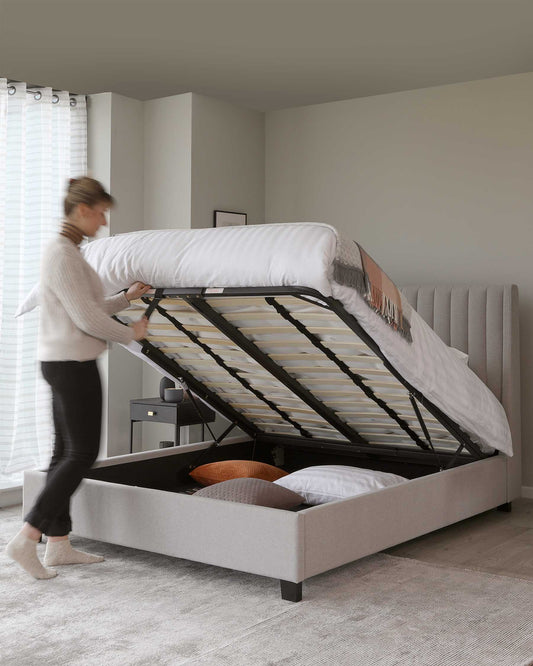 Contemporary light grey upholstered queen storage bed with a lifted mattress base revealing under-bed storage containing pillows and blankets; featuring a tufted headboard and wooden slats, supported by black block legs.