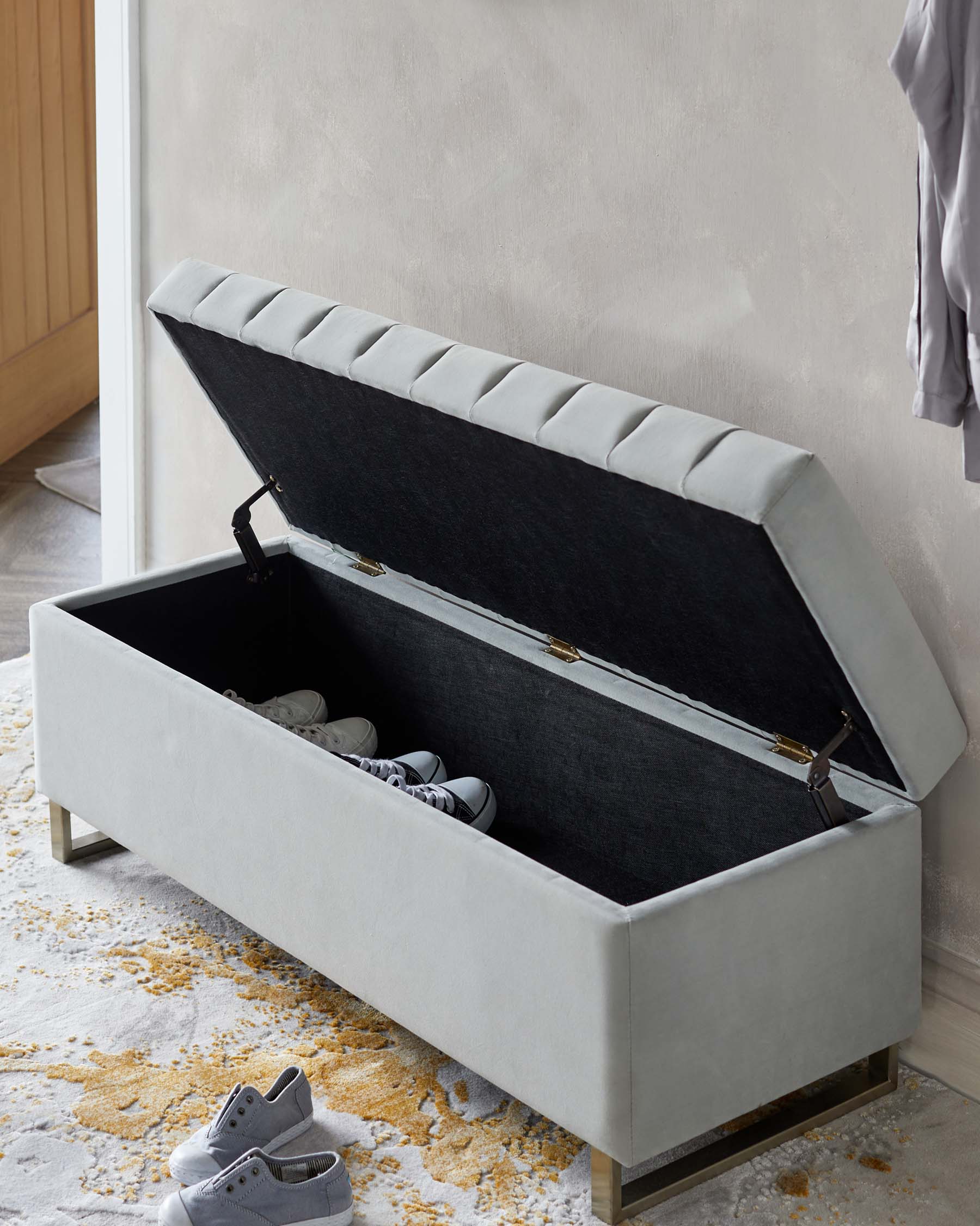 A modern light grey upholstered storage bench with a tufted cushioned lid that is open, revealing a black interior with several pairs of shoes inside. The bench is placed on a patterned rug and features metal accents on the legs.