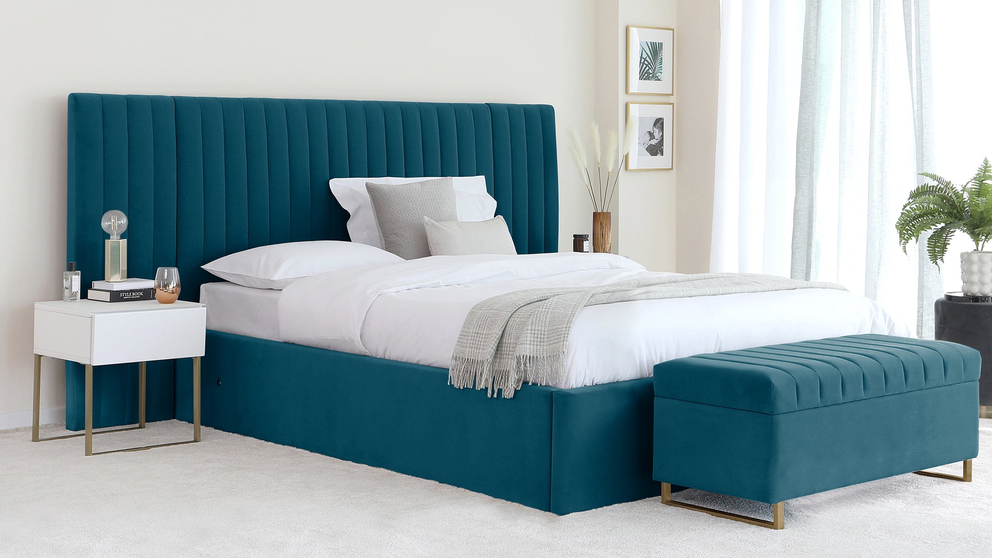 Elegant bedroom furniture featuring a plush teal upholstered bed with an extra tall, channel-tufted headboard, and a matching bench at the foot of the bed. Beside the bed is a minimalist white nightstand with gold metal legs.